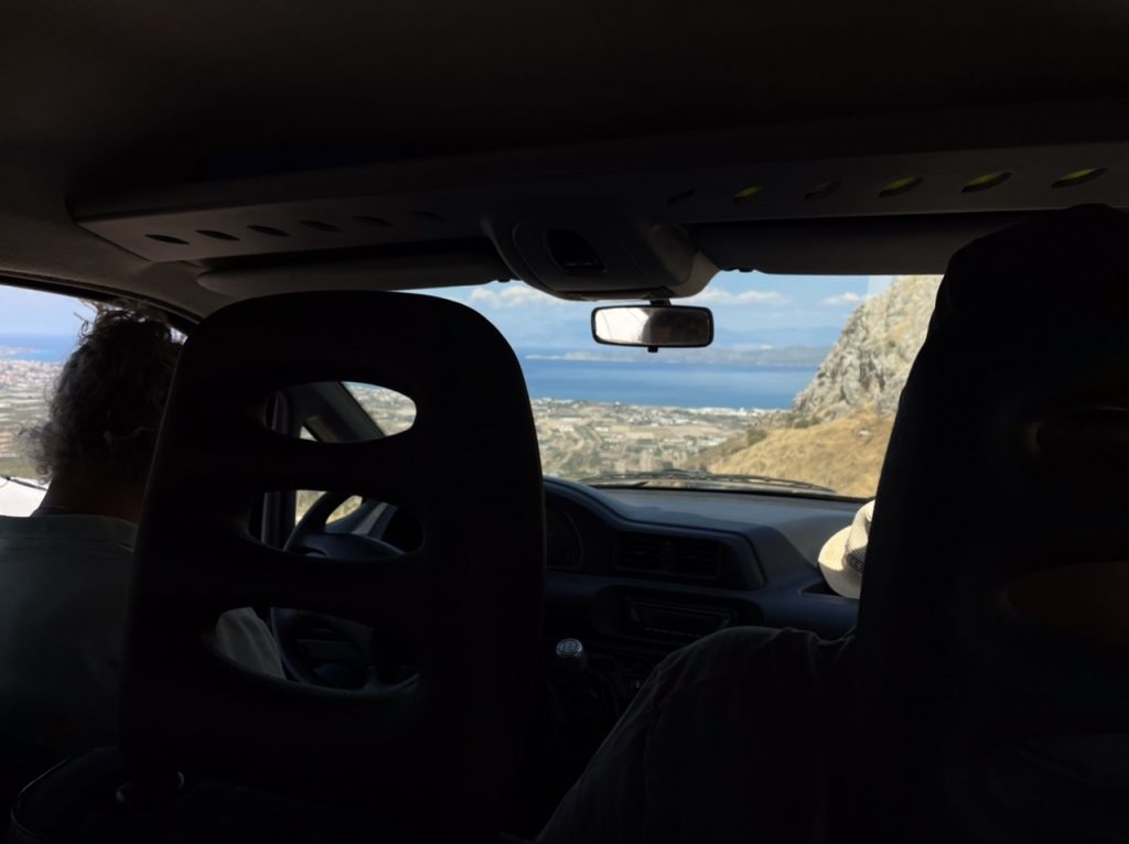 Blick aus einem Auto durch die Frontscheibe auf Felder und Gebäude von einem Berg herab. Im Hintergrund Meer.